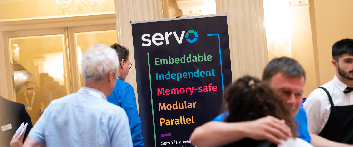 Servo banner in the background showing Servo logo and kewords: embeddable, independent, memory-safe, modular, parallel; with some people around during the Linux Europe Member Summit in Bilbao at 18th September 2023.