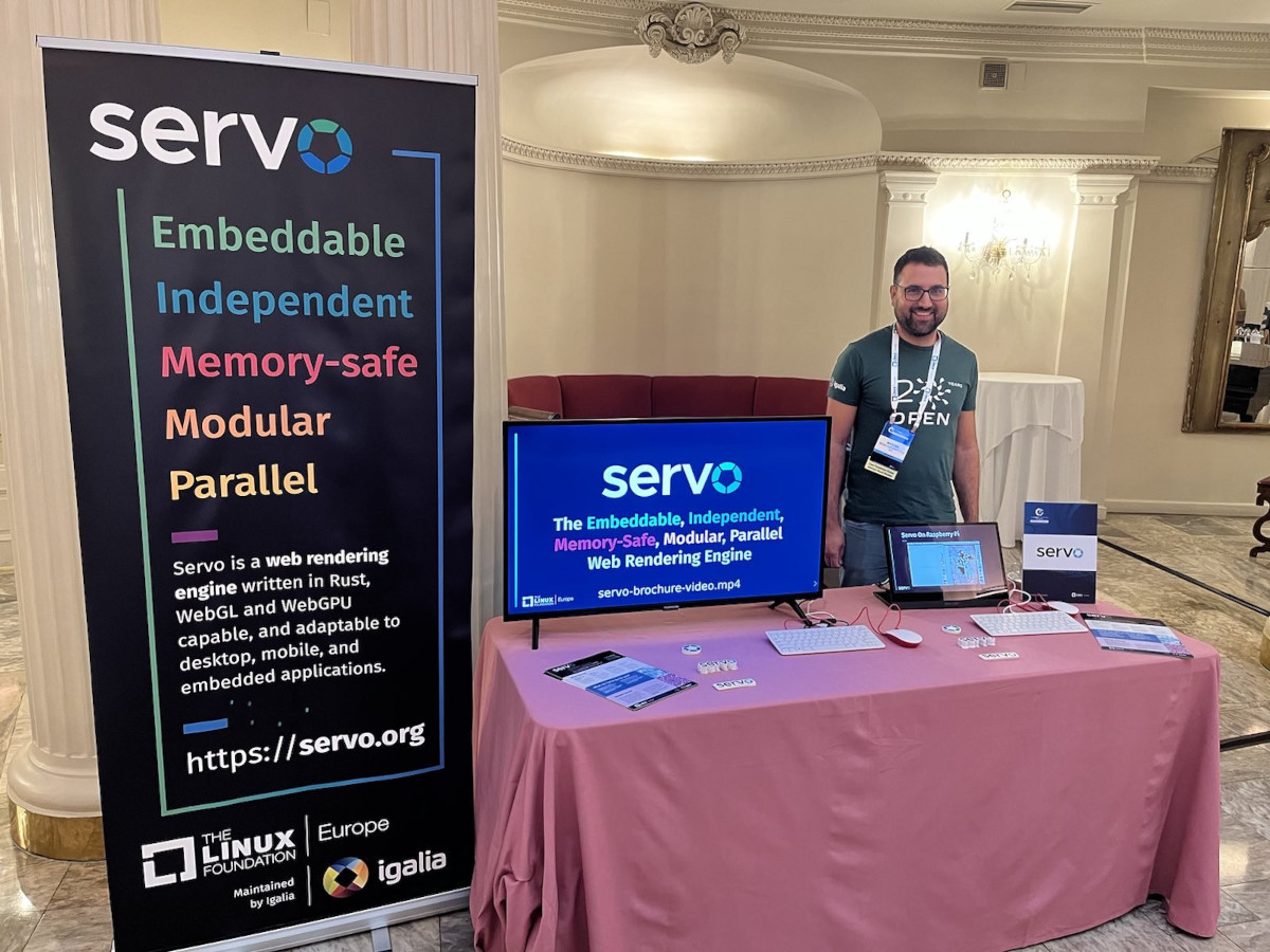 Picture of the Servo booth at Linux Foundation Europe 2023. There's a banner with Servo logo and some brief explanation, then two screens and two Raspberry Pi 400 running some Servo videos. In the booth there are also stickers and brochures about Servo. Manuel Rego is behind the booth.