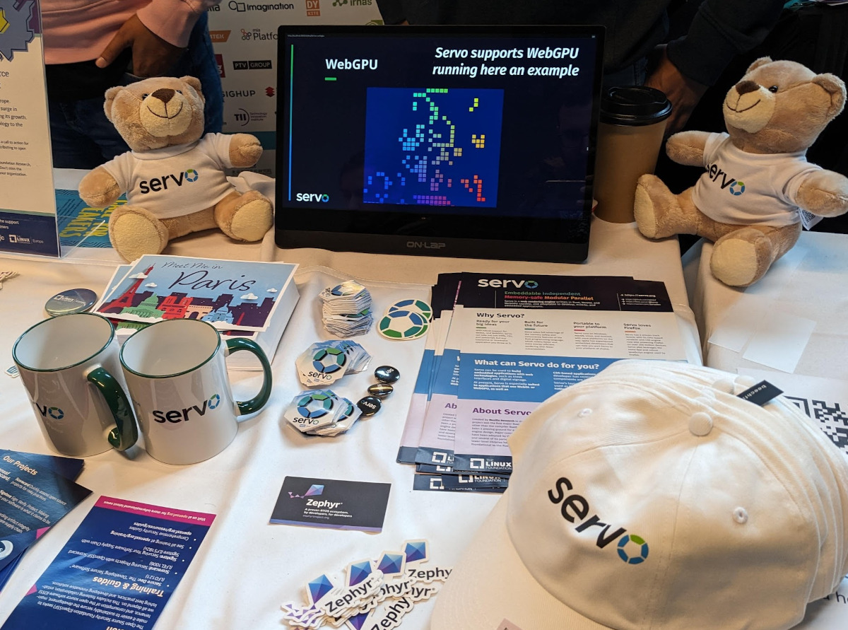 Picture of Linux Foundation Europe stand at FOSDEM 2024 with different Servo swag (stickers, flyers, buttons, cups, caps, teddy bears). Also in the picture there is a screen showing a video of Servo rendering WebGPU content.