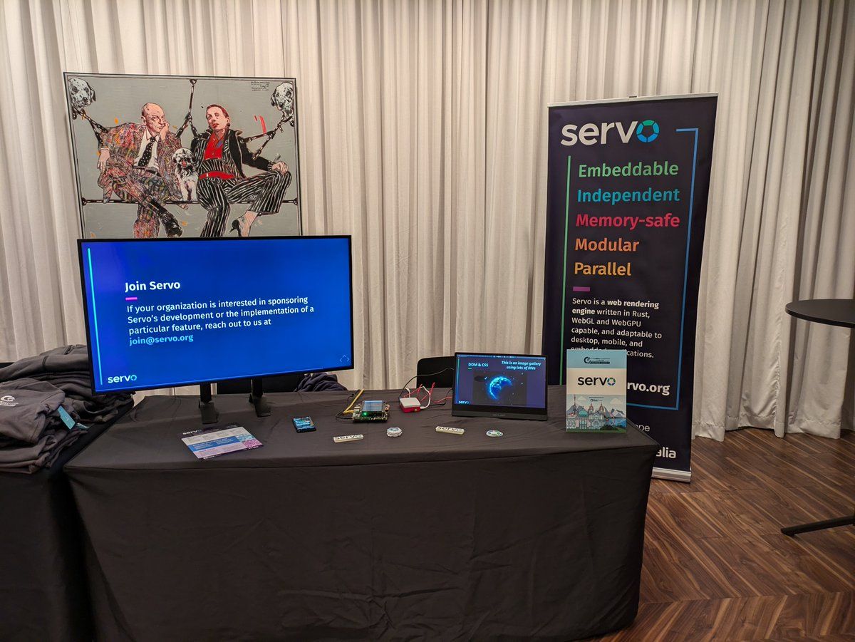 Picture of the Servo booth at Linux Foundation Europe Member Summit 2024 in Vienna. It has a TV showing some Servo videos from YouTube, an Android phone, an Open Harmony board, and a Raspberry Pi 5; where Servo can be showcased. It also has a Servo banner in the background, and several stickers and and flyers in the booth table.