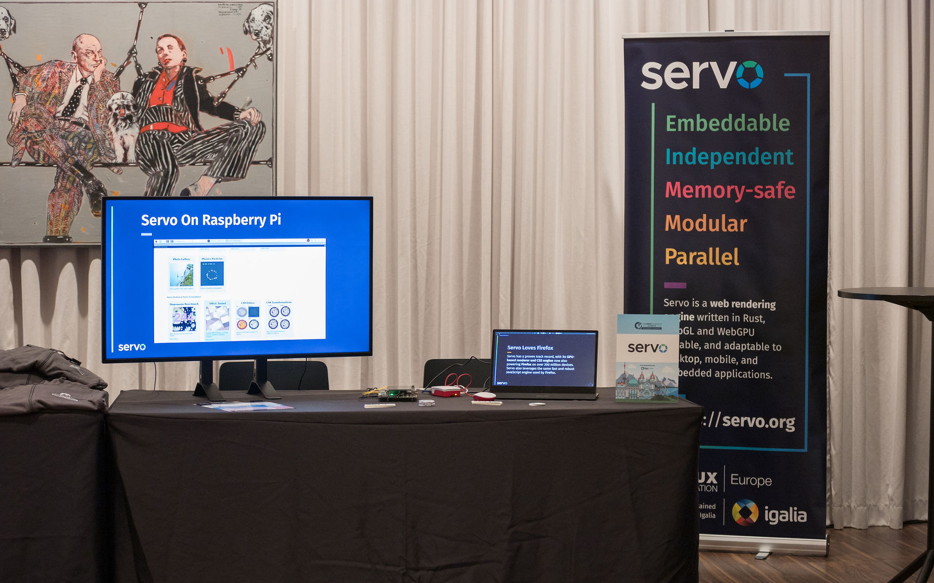 Picture of the Servo booth at Linux Foundation Europe Member Summit 2024. It's a talbe with a TV on the left showing a video of Servo running on Raspberry Pi, there's also a smaller screen on the right, showing a presentation about Servo. Behind the table there's a Servo banner. Over the table there is a Raspberry Pi, a Rockchip board and an Android phone. There are also stikcers and flyers.