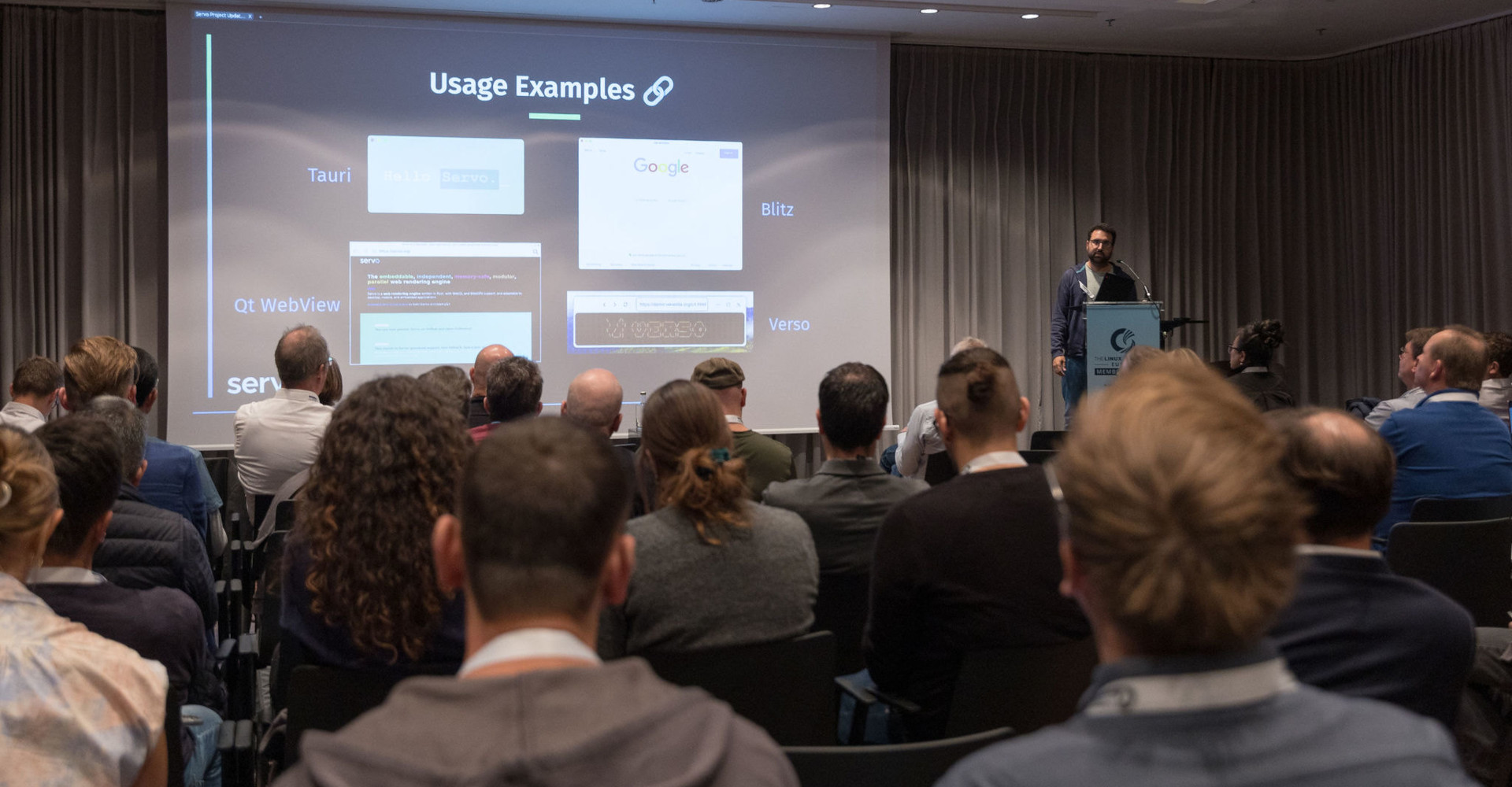 Picture of the Servo Project Updates talk at Linux Foundation Europe Member Summit 2024. In the picture you can see a room with a group of people sitting watching the presentation. On the projector there is a slide about Servo usage exmaples showing Tauri, Qt WebView, Blitz and Verso. Manuel Rego is giving the talk at the right of the projector.